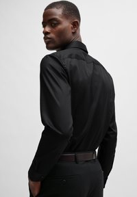 Camisa negra de manga larga con textura suave, con cuello y puños abotonados, combinada con pantalones oscuros y un cinturón marrón.