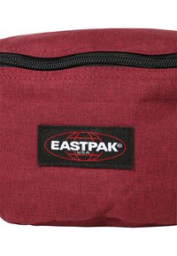 Funda de tela burdeos con una cremallera negra y forma rectangular. Presenta un prominente parche con el logo de Eastpak con acentos rojos.