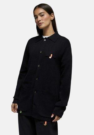 Femme aux longs cheveux foncés portant une veste noire à boutons avec poches avant et détails orange sur les languettes, debout devant un fond clair uni.