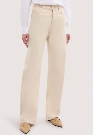 Personne portant un pantalon crème taille haute à jambes larges, associé à des chaussures beiges pointues et une chemise blanche à manches longues, debout sur un fond neutre.