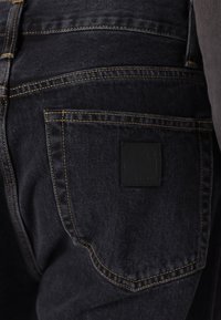 Los pantalones vaqueros de mezclilla negra presentan costuras doradas, un bolsillo trasero con una etiqueta negra rectangular de la marca y una textura suave.