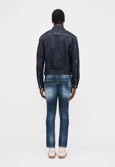 Hombre de pie de espaldas, con una chaqueta de mezclilla oscura y vaqueros azules descoloridos, calzando zapatillas blancas, sobre un fondo liso y claro.