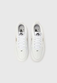 Τα λευκά αθλητικά παπούτσια Nike Air Force 1 διαθέτουν λείο δέρμα, διάτρητη μύτη, λευκά κορδόνια και μαύρο λογότυπο Nike στο πλάι και τη γλώσσα.