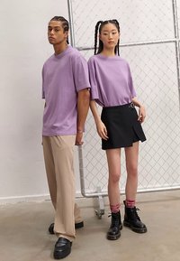 Dwie modelki stoją obok siebie: jedna w luźnym lawendowym t-shircie i beżowych spodniach, druga w lawendowym t-shircie i czarnej spódnicy z paskiem.