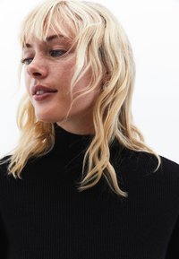 Maglione nero a collo alto a coste con un tessuto texture, design aderente e scollo alto, indossato da una persona con capelli biondi e mossi.