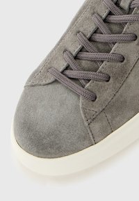 Nærbillede af tåen på en grå ruskindsneaker med matchende snørebånd og hvid gummisål på en hvid baggrund.