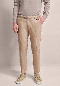Beige maßgeschneiderte Hose mit glatter Textur, ausgestattet mit einer einzigen Falte und einem vorderen Knopfverschluss, kombiniert mit hellen Sneakers.