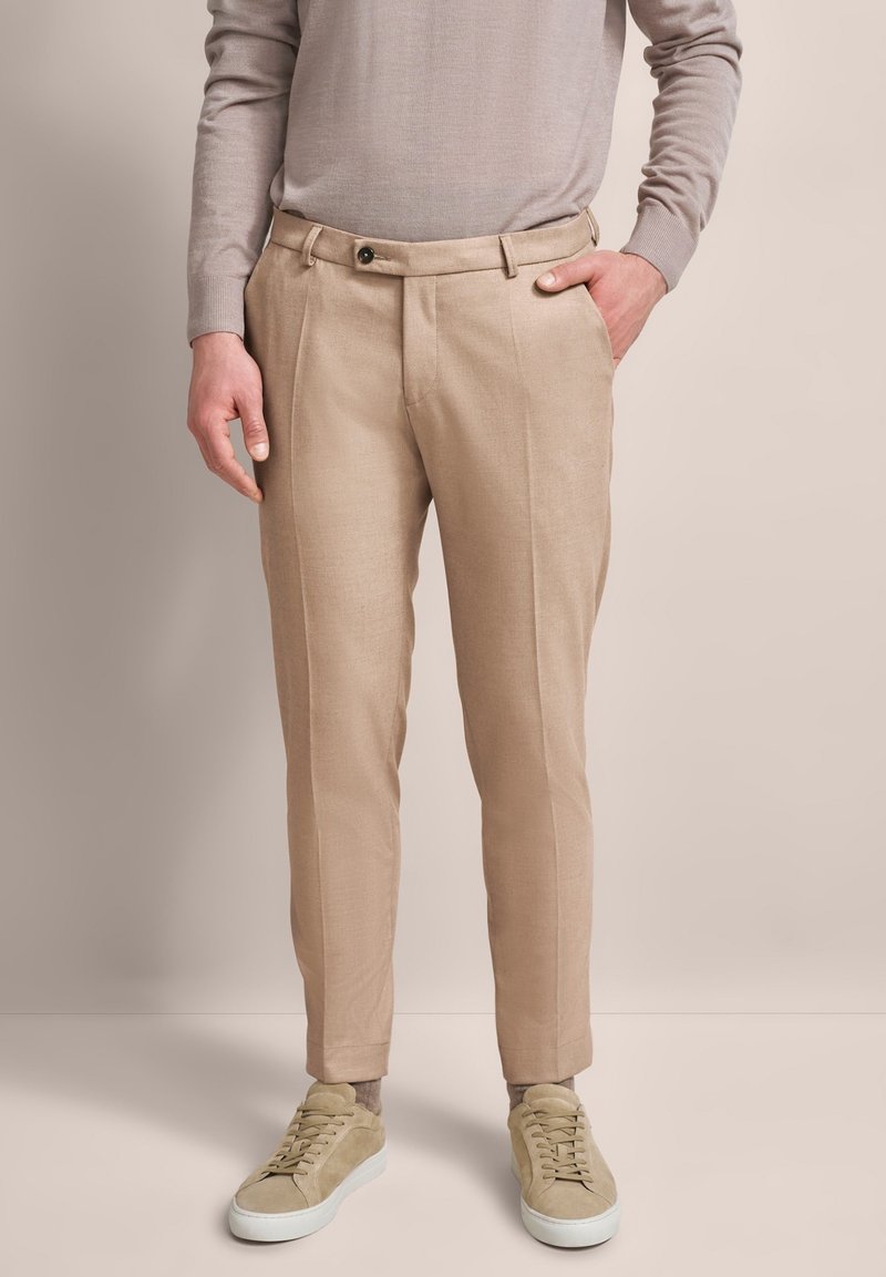 Beige maßgeschneiderte Hose mit glatter Textur, ausgestattet mit einer einzigen Falte und einem vorderen Knopfverschluss, kombiniert mit hellen Sneakers.