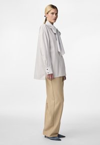Blouse oversize rayée blanche avec un grand col noué, associée à un pantalon beige à jambes larges et des escarpins pointus bleus.