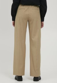Beige weite Hosen mit taillierter Passform, die eine glatte Textur aufweisen, zwei Gesäßtaschen und eine dezente Falte auf der Vorderseite haben.