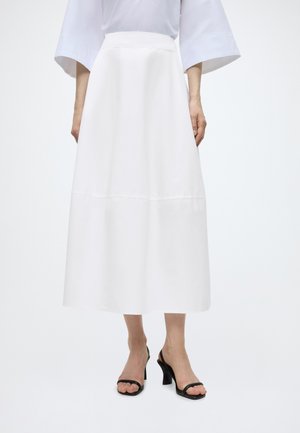 Femme portant une jupe midi blanche taille haute, un chemisier blanc à manches larges et des sandales à talons noires ouvertes aux orteils.