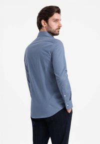 Camisa de manga larga azul claro con un cuello estructurado, tejido texturizado y puños con botones, combinada con pantalones oscuros de cuadros.