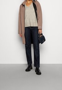 Brązowy wodoodporny płaszcz na jasnoszarym swetrze, ciemne dżinsy i czarne botki za kostkę. Mała granatowa torebka z wzorzystym paskiem.