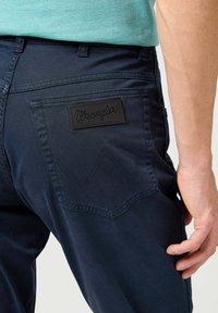Pantalones Wrangler azul marino con una etiqueta negra en el bolsillo trasero, con un patrón de costura prominente y una superficie texturizada.