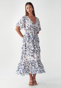 Robe maxi florale blanche avec éclaboussures bleu marine, décolleté en V, manches courtes flottantes, taille cintrée et volants superposés à l'ourlet.