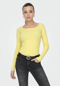 Geel geribbeld langemouwen top met een vierkante halslijn, gecombineerd met hoge zwarte jeans en een zwarte riem met een zilveren dubbele cirkelsluitingen.