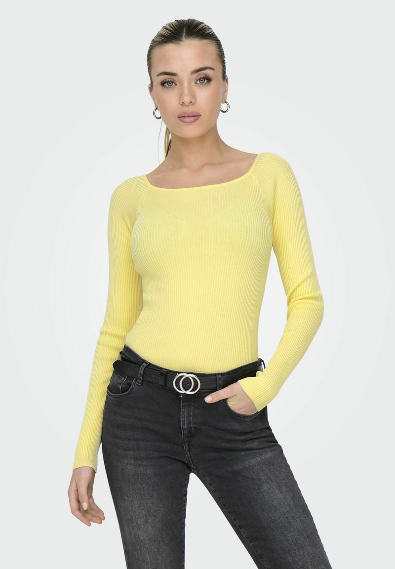 Geel geribbeld langemouwen top met een vierkante halslijn, gecombineerd met hoge zwarte jeans en een zwarte riem met een zilveren dubbele cirkelsluitingen.