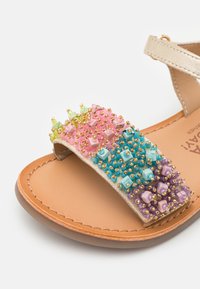 Sandal colorido con una suela plana en tono marrón, una tira ancha adornada con cuentas en tonos pastel de rosa, azul, verde y morado, y detalles dorados.