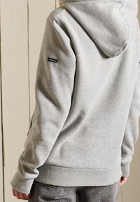 Sudadera con capucha gris hecha de tela suave y cepillada, con capucha ajustable con cordón y puños acanalados. Incluye un parche con logo en el hombro izquierdo.