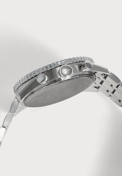 Montre-bracelet en métal argenté vue de côté montrant une couronne gravée "BOSS" et deux boutons-poussoirs sur un bracelet à maillons sur fond uni.
