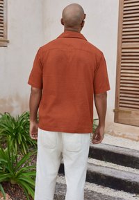 Rust-colored short-sleeve shirt with sunburst embroidery, paired with white pants. Features a standard collar and a relaxed fit.
