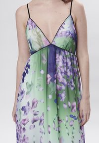 Abito maxi floreale con scollatura profonda, caratterizzato da motivi di fiori verdi e viola, sottili spalline nere e una silhouette fluida.