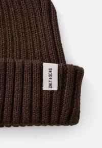Brown knit beanie with ribbed texture. Features a white tag with "ONLY" and "SHOES" printed. Soft, flexible fabric designed for warmth.