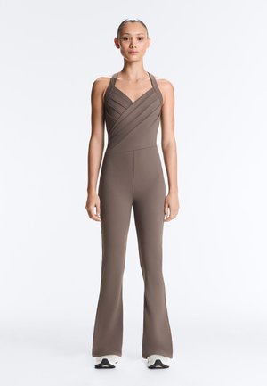 Vrouw staand, voor zich uit kijkend, gekleed in een nauwsluitende taupekleurige mouwloze jumpsuit met gekruiste borstdetails en witte sneakers.