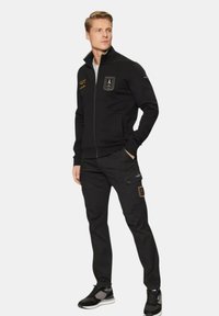 Zwarte zip-up jas met patches, gecombineerd met bijpassende zwarte broek. De outfit heeft een slim fit ontwerp en een soepele stof. Ontworpen voor comfort.