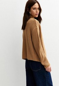 Maglione a maglia waffle color marrone chiaro, vestibilità rilassata, maniche lunghe. Abbinato a jeans a gamba larga blu scuro, che mettono in mostra un tessuto testurizzato e un design informale.