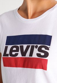 Camiseta de algodón blanca con un gran logo negro de "Levi's" y acentos azules y rojos en franjas horizontales. Cuello redondo simple, mangas cortas.