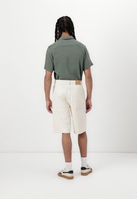 Short-sleeve green shirt with a straight hem, paired with cream-colored shorts. Wearer has braided hair, shows back view, with neutral background.