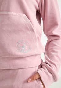 Vaaleanpunainen huppari, jossa on strasseilla varustettu "GL" monogrammi taskussa, jota käyttää henkilö, jonka käsi on osittain työnnettynä yhteen myöhemmin sopiviin vaaleanpunaisiin housuihin.