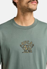 T-shirt in cotone verde con logo a stampa leopardata in nero e giallo senape. Collo a giro classico con maniche corte.