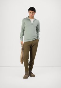 Suéter verde claro con cremallera, pantalones marrones y botas marrones con cordones. El modelo sostiene un bolso beige y está de pie sobre un fondo liso.