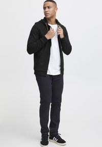 Sudadera negra con cremallera y cordón, combinada con una camiseta blanca y pantalones oscuros azul marino. Zapatillas negras con rayas blancas. Diseño sencillo.