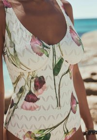 Femeie purtând un costum de baie alb cu modele florale lângă plajă, cu mare albastră și țărm stâncos în fundal.