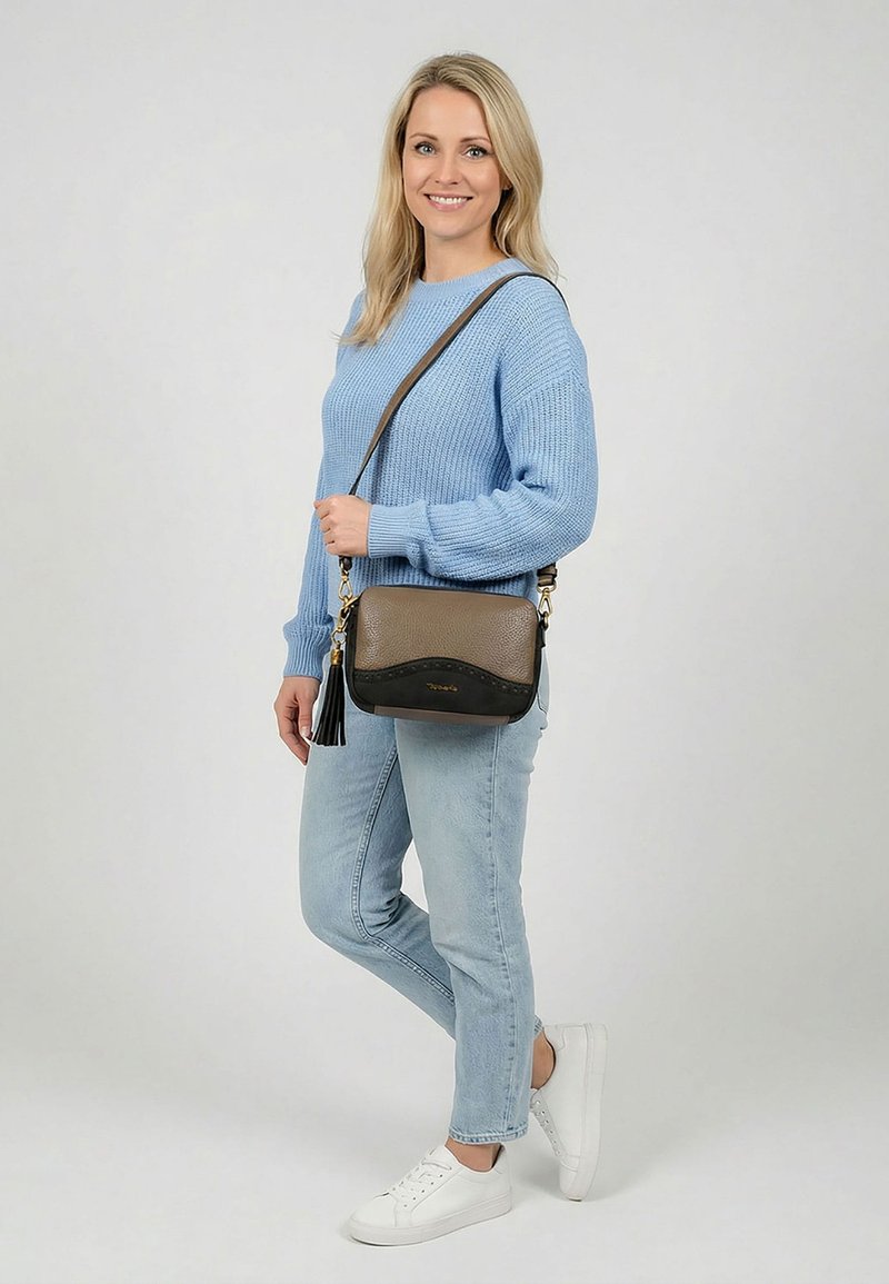 Femme souriante, portant un pull bleu clair, un jean clair, des baskets blanches et un sac bandoulière marron avec un détail à pampilles.