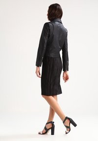 Giacca di pelle nera con un design aderente, abbinata a una gonna nera plissettata. Il modello indossa scarpe con tacchi alti neri e strappy.