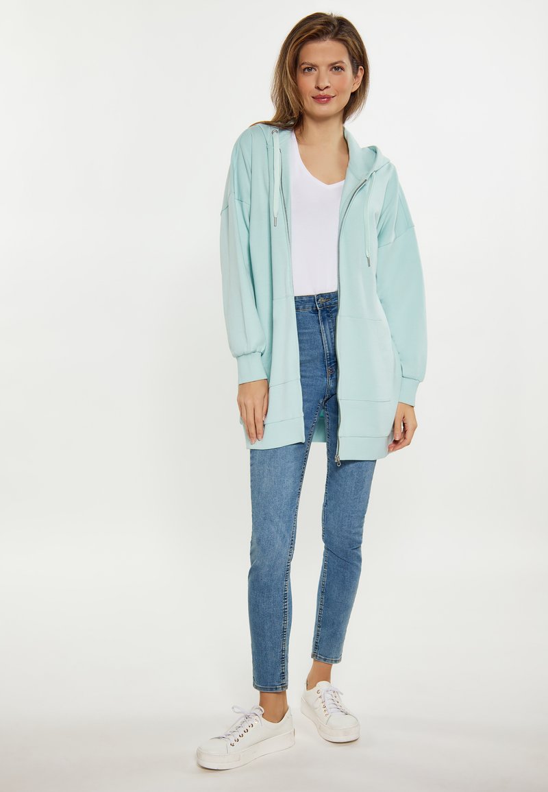 Felpa oversize verde menta con cerniera, cordini e tasche laterali, abbinata a jeans skinny blu chiaro e scarpe da ginnastica bianche.