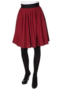 Gonna plissettata rossa con una vita nera, lunghezza al ginocchio, caratterizzata da una texture di tessuto liscia. Abbinata a collant neri e tacchi alti.