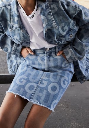 Persona que lleva un conjunto de chaqueta vaquera y falda con grandes logotipos "HUGO", combinado con una camiseta blanca que también lleva la marca "HUGO" en el escote.
