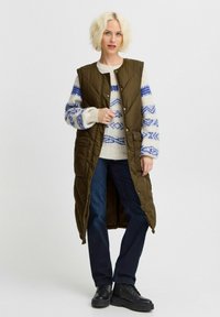 Olivengrüne, lange Pufferweste mit einem gesteppten Design, kombiniert mit einem cremefarbenen Pullover mit blauen geometrischen Mustern und dunklen Jeans.