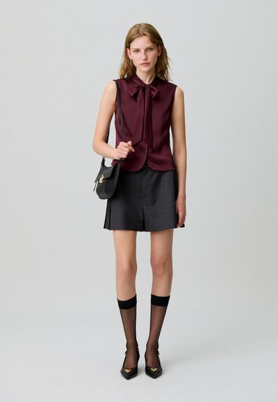 Blusa senza maniche bordeaux con dettaglio a fiocco, abbinata a pantaloni corti plissettati grigio scuro. Gli accessori includono una borsa a spalla nera e calze fino al ginocchio.