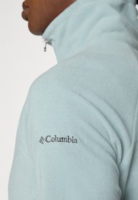 Giacca in pile azzurro chiaro con colletto alto e zip al collo. Presenta un logo Columbia grigio scuro sulla spalla sinistra. Texture morbida.