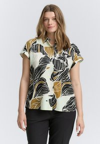 Korte mouwen blouse in lichtgroene stof met een bladerenpatroon in zwart en mosterd. Heeft een knoopsluiting aan de voorkant en een relaxte pasvorm.