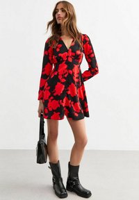 Schwarzes Kleid mit rotem Blumenmuster, langen Ärmeln, V-Ausschnitt, ausgestelltem Rock, kombiniert mit schwarzen Ankle Boots und einer schwarzen Handtasche.