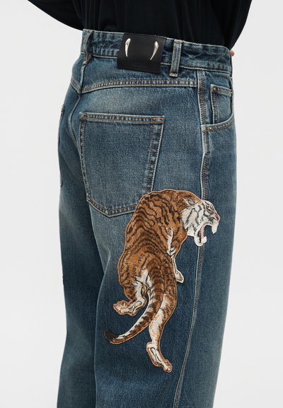 Σκούρο μπλε τζιν με κεντημένη τίγρη που ουρλιάζει στο μηρό και μαύρο δερμάτινο έμπλαστρο με ασημένιες γρατζουνιές νυχιών στη μέση.