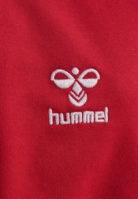 Rote Stoff mit einer glatten Textur, der ein white gesticktes Logo von "hummel" zeigt, das ein stilisiertes Design einer Biene oder eines Schmetterlings darstellt.