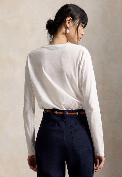 Γυναίκα με μαύρα μαλλιά δεμένα πίσω, φοράει λευκό πουκάμισο με μακριά μανίκια, σκούρο μπλε παντελόνι, καφέ ζώνη και σκουλαρίκια σε σχήμα κοχυλιού, στρέφοντας το πρόσωπο πλευρικά.